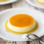 A straight-on view of homemade flan that has a light caramel sauce on top of it and surrounding the base and is on a white plate with other plated servings nearby.