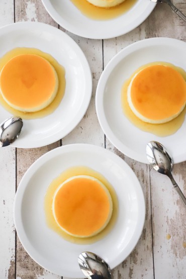 An overhead view of four white dessert plates that are holding homemade flan that are resting in a small pool of a light caramel sauce.