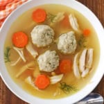 An overhead view of a shallow white soup bowl filled with homemade matzo ball soup with chicken and carrots and several sprigs of fresh dill.