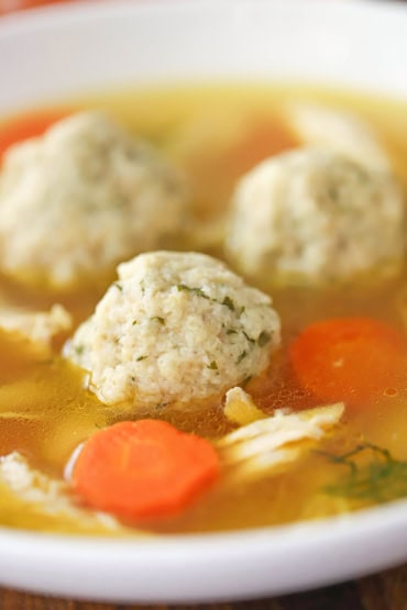 A close-up view of a three homemade matzo balls in a soup bowl of chicken broth a pieces of chicken and carrot slices.
