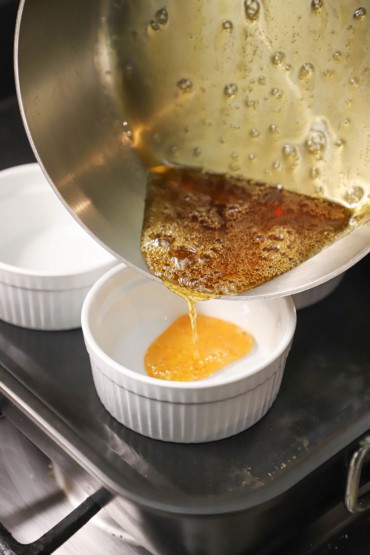 A person pouring a light caramel sauce from a silver saucepan into an 8-ounce ramekin that is sitting in a roasting pan partially filled with water.