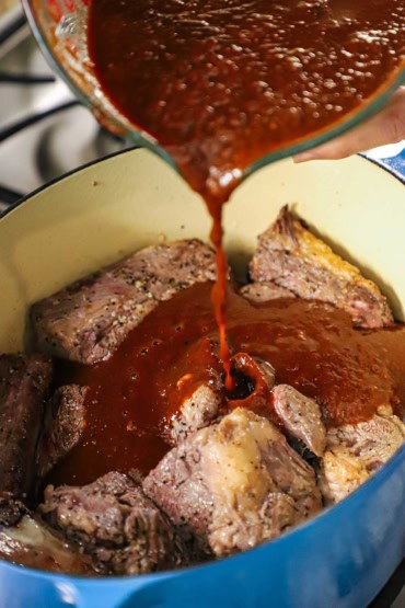 A person pouring an ancho pepper sauce into a large oval Dutch oven that is filled with chunks of seared beef chuck roast and short ribs.