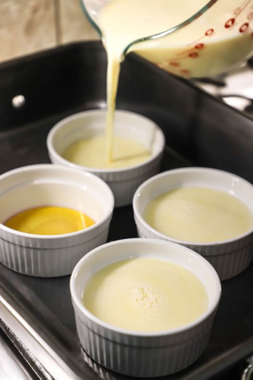 A person pouring a vanilla custard into 8-ounce ramekins that are sitting in a large roasting pan that is partially filled with water.