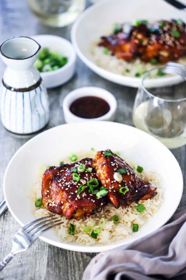 A straight-on view of two white dinner bowls filled with a serving of baked teriyaki chicken resting on a bed of steamed rice with a glass of white wine near by.