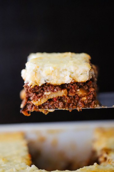 A piece of moussaka that consists of a meat sauce, potatoes, eggplant, and a thick bechamel being raised from a baking dish on a metal spatula.
