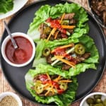 An overhead view of three beef bulgogi lettuce wraps that are sitting on a black plate and next to a small bowl filled with Korean barbecue sauce.