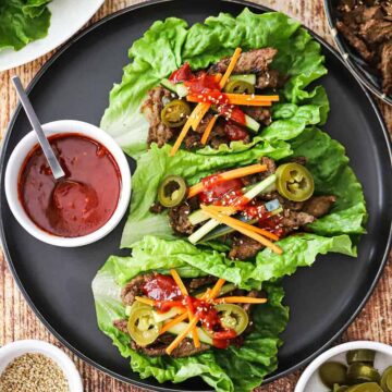 An overhead view of three beef bulgogi lettuce wraps that are sitting on a black plate and next to a small bowl filled with Korean barbecue sauce.
