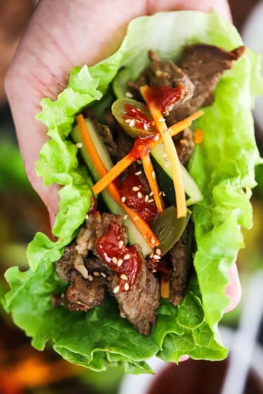 A person holding Korean beef bulgogi lettuce wrap that is topped with julienned carrots and cucumber as well as pickled peppers and sesame seeds.