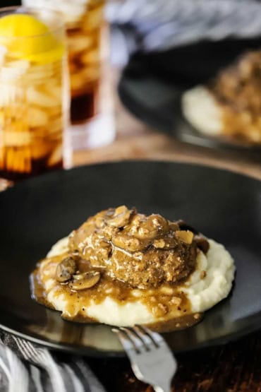 A straight-on view of a salisbury steak with mushroom brown gravy sitting in the middle of a black dinner plate next to two glasses of iced tea with lemon.