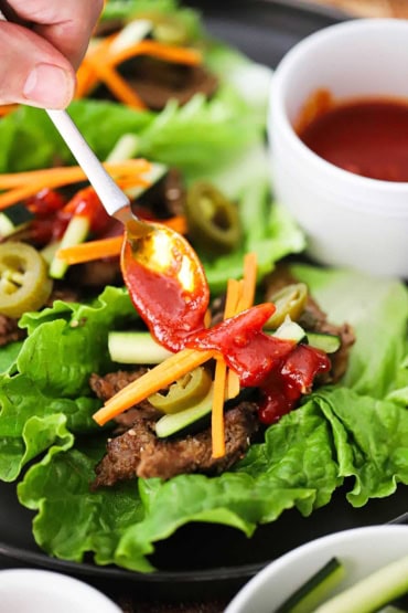 A person using a small spoon to drizzle Korean barbecue sauce over strips of bulgogi beef nestled into a lettuce leaf and toped with julienned carrots and cucumber.