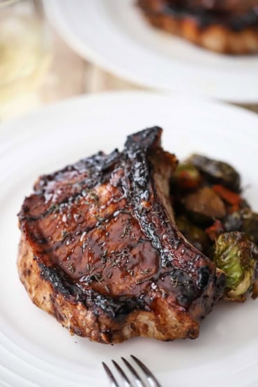 A close-up view of a grilled balsamic glazed pork chop that is resting on a pile of roasted Brussels sprouts and carrots on a white dinner plate.