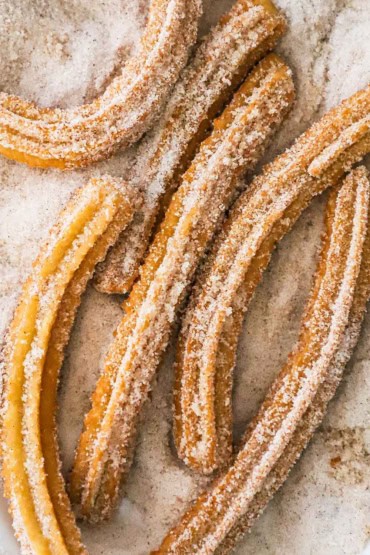 An overhead view of a homemade churros that are sitting on a platter that is filled with a sugar and cinnamon mixture.