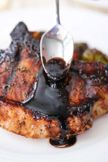 A person using a small spoon to drizzle a balsamic glaze or a grilled pork chop that is resting on a white dinner plate.