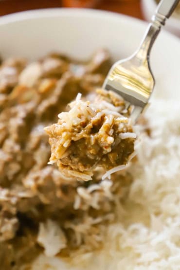 A person using a silver fork to lift up a helping of chicken korma mixed with basmati rice over a bowl of the same.