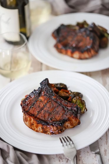 A straight-on view of a grilled balsamic glazed pork chop that is sitting next to a pile of roasted vegetables with another plate of the same in the background.