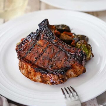 A straight-on view of a grilled balsamic glazed pork chop that is sitting next to a pile of roasted vegetables.