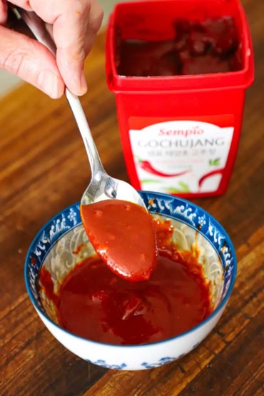 A person holding a spoonful of Korean barbecue sauce over a small bowl of the same with a container of guchujang next to it.