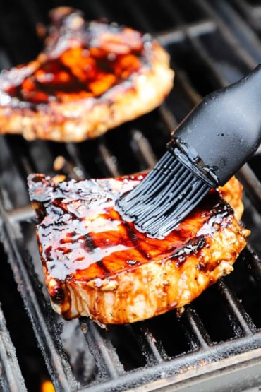 A person using a grill brush to apply a thick balsamic glaze over the top of a center-cut pork chop that is being grilled on a gas grill.