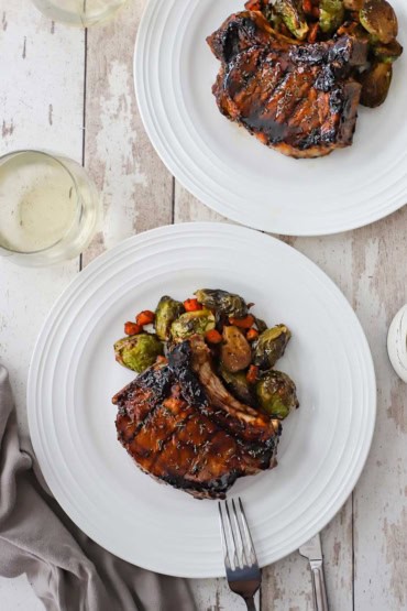 An overhead view of two white dinner plates filled with serving of grilled balsamic glazed pork chops with roasted Brussels sprouts and carrots.