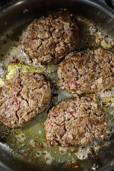 A close-up view of four partially cooked salisbury steaks that are simmering in butter and oil in a large silver skillet.