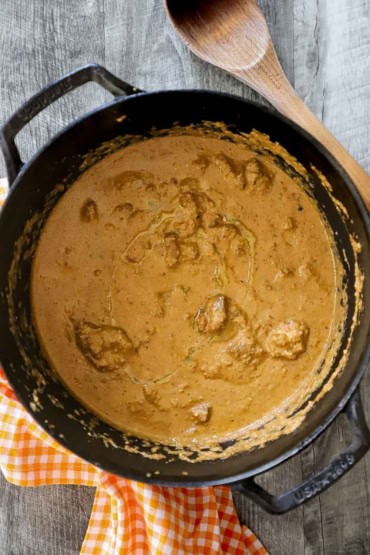 An overhead view of a black pot filled with chicken korma with a orange checkered napkin and wooden spoon nearby.