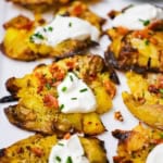 A close-up view of cooked smashed potatoes that are brown and crispy on the edges that have been topped with crumbled bacon, a dollop of sour cream, and snipped chives.