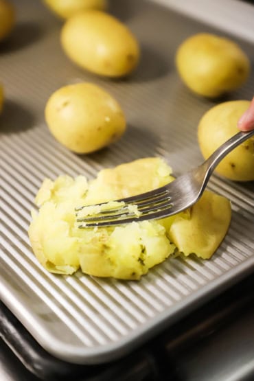 A close-up view of a person using a fork to smash a tender gold potato on a metal baking pan.
