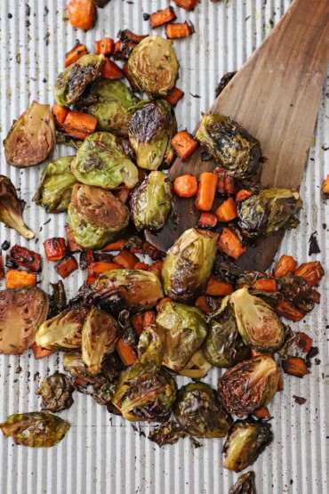 An overhead view of halved Brussels sprouts and pieces of carrots that have been tossed with balsamic vinegar and herbs and roasted on a large silver baking sheet.