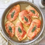 An overhead view of six fully cooked chicken saltimbocca cutlets in a skillet that are topped with crispy prosciutto and a sage leaf all resting in a white wine butter sauce.