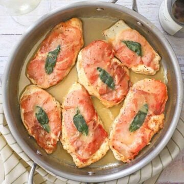 An overhead view of six fully cooked chicken saltimbocca cutlets in a skillet that are topped with crispy prosciutto and a sage leaf all resting in a white wine butter sauce.