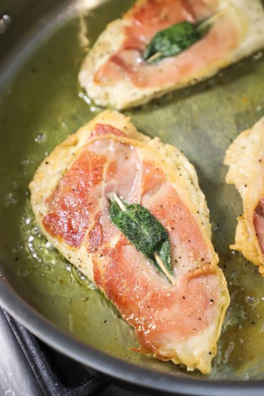A close-up view of a thin chicken cutlet that has been seared in a skillet with oil and butter and is topped with a strip of prosciutto and a fresh sage leaf.