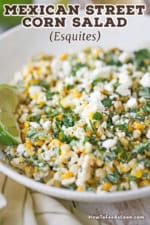A close-up view of a white bowl filled with Mexican street corn salad with a lime wedge resting on the side and garnished with chopped parsley and crumbled Cotija cheese.