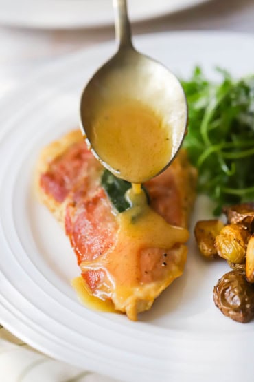 A large silver spoon being used to drizzle a white wine butter sauce over the top of a chicken saltimbocca cutlet next to roasted potatoes and an arugula salad.