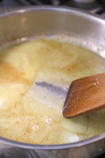 A close-up view of a large silver saucepan filled with a white wine butter sauce that is being stirred with a wooden spatula.