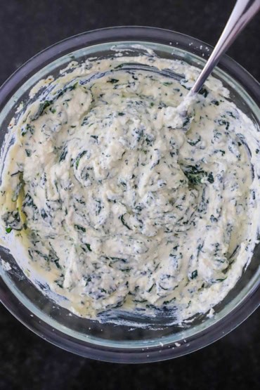An overhead view of a large glass bowl that is filled with a ricotta and chopped spinach mixture with a large silver spoon inserted off to the side of the bowl.
