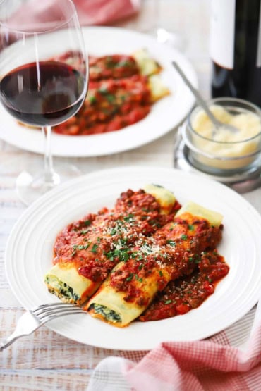 A straight-on view of a white dinner plate that is filled with two side-by-side spinach cannelloni and meat sauce near a wine glass partially filled with red wine.