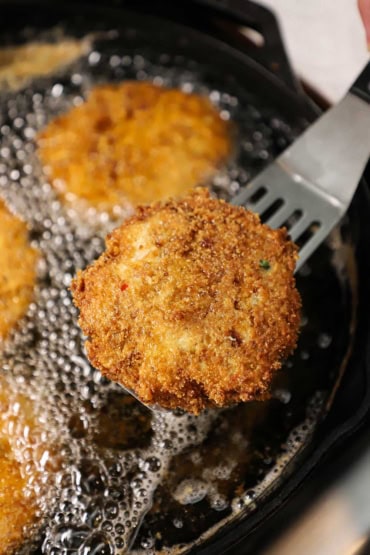 A person holding a metal spatula that has a crispy crab cake on it that is being lifted from a cast-iron skillet filled with hot oil.