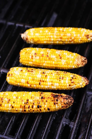 A close-up view of four ears of fresh sweet corn being lightly charred on a gas grill.