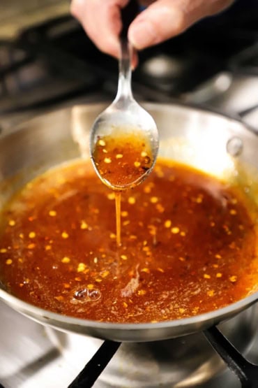 A person lifting up a silver dinner spoon filled with hot honey sauce of a small skillet filled with the same.