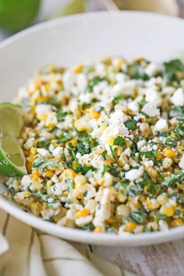 A close-up view of a white bowl filled with Mexican street corn salad with a lime wedge resting on the side and garnished with chopped parsley and crumbled Cotija cheese.