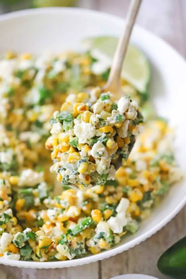 A close-up view of a large serving spoon lifting up a helping of Mexican street corn salad over a bowl filled with the same.