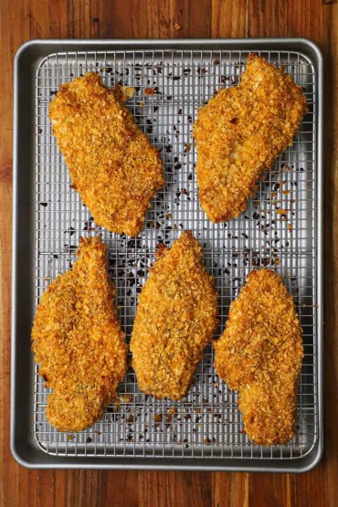 An overhead view of five baked corn-flake encrusted chicken cutlets all resting on a baking rack in a baking pan.