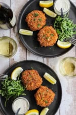 An overhead view of two black dinner plates that are each holding two crispy New England-style crab cakes next to lemon wedges, a jar of tartar sauce, and an arugula salad.