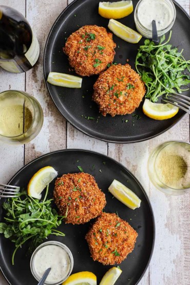 An overhead view of two black dinner plates that are each holding two crispy New England-style crab cakes next to lemon wedges, a jar of tartar sauce, and an arugula salad.
