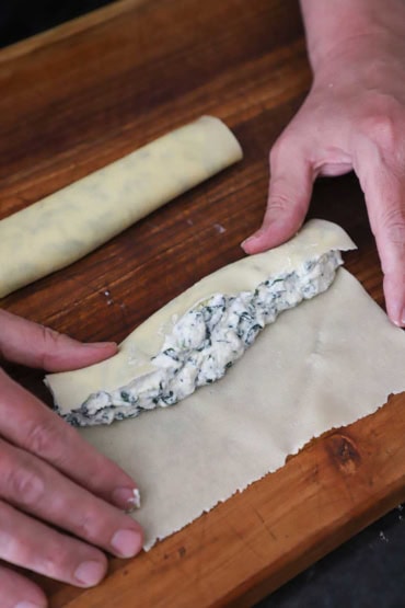 A person rolling a square piece of freshly made egg pasta around a full-length tube of a ricotta and spinach filling.