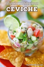 A close-up view of a small glass bowl that is filled with a serving of Mexican shrimp ceviche and is on a green cocktail plate with corn tortilla chips and a silver spoon on it.
