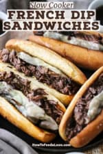 A straight-on view of four large slow cooker French dip sandwiches resting on a black plate with two small bowls filled with au jus sauce nearby.