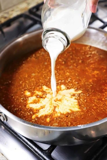 A person pouring half and half from a glass milk jug into a large skillet filled with a simmering red meat sauce.