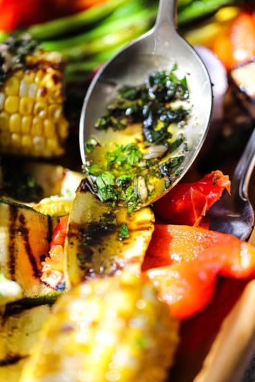 A person using a silver serving spoon to drizzle Italian dressing over grilled vegetables on a bamboo platter.