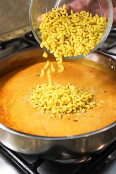 A person dumping uncooked macaroni elbows from a glass bowl into a skillet filled with a creamy tomato and beef sauce.